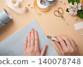 Hands cut off a piece of pastel polka dot paper. Ribbons, scissors, flowers and craft supplies on beige background 140078748