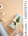 Vertical flat lay of light blue polka dot gift box tied with cream ribbon and thuja twig. Vintage scissors, pearls, and wrapping supplies on a beige background. Copyspace 140078751