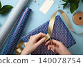 Hands cutting a gold ribbon with scissors on a polka dot wrapped gift box. Wrapping paper rolls and crafting accessories on a blue background. 140078754