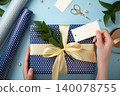 Hands are attaching tag to gift box wrapped in blue polka-dot paper, tied with a bow made of gold satin ribbon. Green leaves, pearls and scissors against a blue background. 140078755