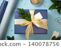 Blue polka dot gift box with a large cream ribbon bow and green twig, surrounded by wrapping paper rolls, scissors, and craft supplies. 140078756