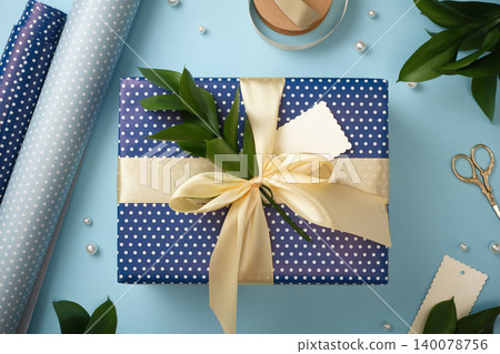Blue polka dot gift box with a large cream ribbon bow and green twig, surrounded by wrapping paper rolls, scissors, and craft supplies. Blue polka dot gift box with a large cream ribbon bow and green twig, surrounded by wrapping paper rolls, scissors, and craft supplies. 140078756