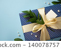 Blue gift box with cream satin bow, green leaves and blank tag on blue background. Space for your text 140078757