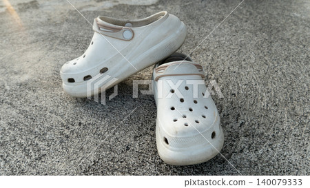Pair of casual white rubber clog sandals placed loosely on a rough concrete outdoor floor. Capturing a simple, everyday footwear scene illuminated by soft sunlight. Pair of casual white rubber clog sandals placed loosely on a rough concrete outdoor floor. Capturing a simple, everyday footwear scene illuminated by soft sunlight. 140079333