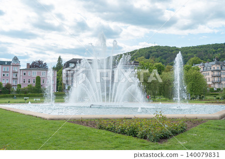 A beautiful public square fountain spraying water high against a background of classic European city apartments and distant green forested hills. Scenic urban landscape under a cloudy summer sky A beautiful public square fountain spraying water high against a background of classic European city apartments and distant green forested hills. Scenic urban landscape under a cloudy summer sky 140079831