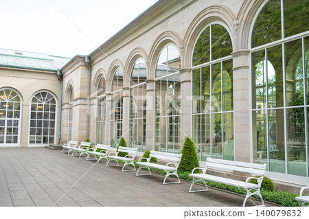 A perspective look at a long grand glass window facade of a historic European pavilion building. A neat row of empty white wooden benches and small trimmed trees standing on a paved walkway. Classical A perspective look at a long grand glass window facade of a historic European pavilion building. A neat row of empty white wooden benches and small trimmed trees standing on a paved walkway. Classical 140079832