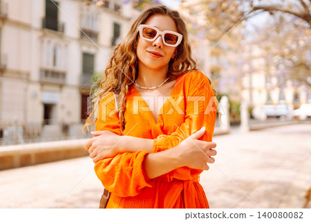 Beautiful young woman in an orange dress enjoys a sunny day in a lively setting with blooming trees Beautiful young woman in an orange dress enjoys a sunny day in a lively setting with blooming trees 140080082