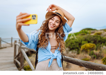 Young woman with curly hair wears a stylish hat and waves while taking a selfie. Travel, tourism Young woman with curly hair wears a stylish hat and waves while taking a selfie. Travel, tourism 140080091