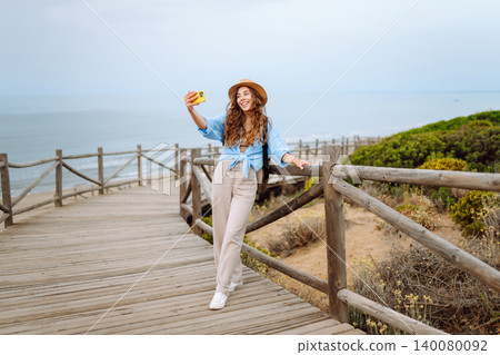 Young woman with curly hair wears a stylish hat and waves while taking a selfie. Travel, tourism Young woman with curly hair wears a stylish hat and waves while taking a selfie. Travel, tourism 140080092
