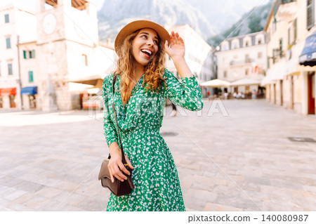 Young woman with curly hair playfully adjusts her wide-brimmed hat while strolling through a square 140080987