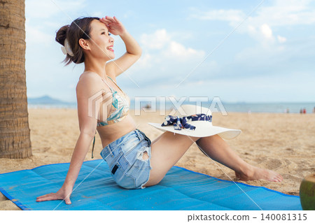 Happy Asian woman sitting on beach mat enjoying summer vacation Happy Asian woman sitting on beach mat enjoying summer vacation 140081315