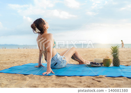 Asian woman relaxing on beach mat with tropical fruits at sunset Asian woman relaxing on beach mat with tropical fruits at sunset 140081380