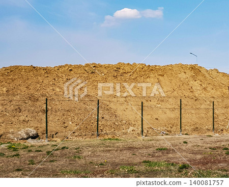 Outdoor construction earthworks with a large pile of sand behind the fence and a heavenly landscape 140081757