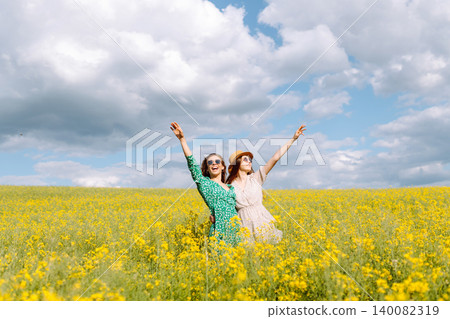 Two young happy women in middle of a yellow rapeseed field. Concept of fun, relaxation. Lifestyle. Two young happy women in middle of a yellow rapeseed field. Concept of fun, relaxation. Lifestyle. 140082319