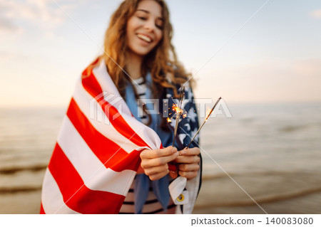 Young woman with American flag and sparklers on beach. Patriotic holiday. USA celebrate 4th of July. Young woman with American flag and sparklers on beach. Patriotic holiday. USA celebrate 4th of July. 140083080