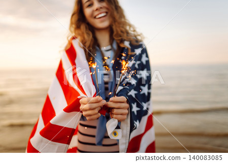 Young woman with American flag and sparklers on beach. Patriotic holiday. USA celebrate 4th of July. 140083085