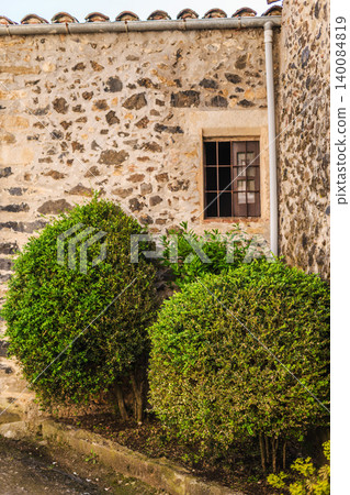 Stone house facade with small wooden window and neatly trimmed green shrubs in traditional Catalan village 140084819