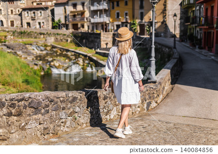Woman in white dress walking along riverside street in historic Catalan village, exploring traditional stone architecture Woman in white dress walking along riverside street in historic Catalan village, exploring traditional stone architecture 140084856