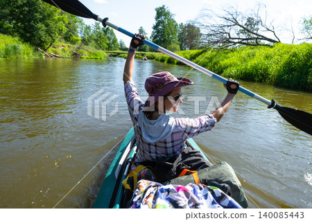 Woman raises paddle above head while kayaking, river trip, active water sport, summer adventure, energetic outdoor lifestyle, tourist use. Woman raises paddle above head while kayaking, river trip, active water sport, summer adventure, energetic outdoor lifestyle, tourist use. 140085443