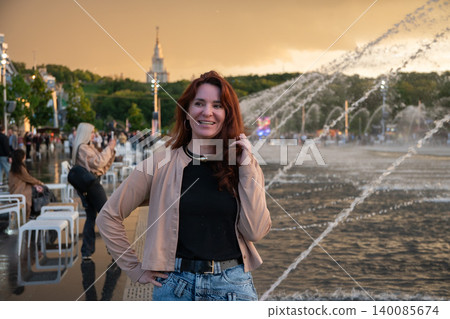 Athletic woman with red hair stands near fountain at Luzhniki embankment, wearing casual clothing, urban waterfront with water jets and city park scenery. Urban recreation promotion, tourism content Athletic woman with red hair stands near fountain at Luzhniki embankment, wearing casual clothing, urban waterfront with water jets and city park scenery. Urban recreation promotion, tourism content 140085674