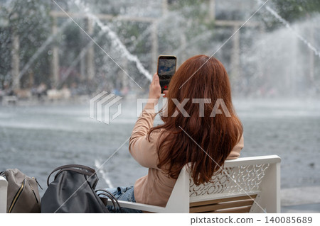 Red-haired woman sits on bench in Luzhniki, photographs fountain with smartphone, water jets, modern urban surroundings creating contemporary travel. Mobile technology, urban leisure 140085689