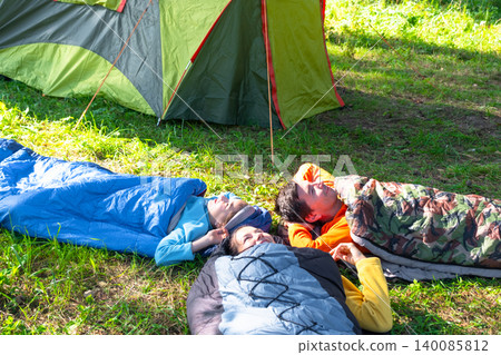 Family camping outdoors: mother, father and daughter resting together in sleeping bags near the tent. Family camping outdoors: mother, father and daughter resting together in sleeping bags near the tent. 140085812