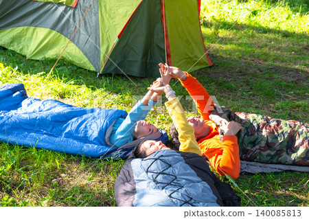 Mother, father and daughter on summer family camping trip, relaxing in sleeping bags near a tent in the forest. Mother, father and daughter on summer family camping trip, relaxing in sleeping bags near a tent in the forest. 140085813