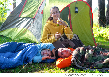 Mother and daughter outdoors by sleeping bags, father close, family camping picnic, healthy gear and sports equipment. Mother and daughter outdoors by sleeping bags, father close, family camping picnic, healthy gear and sports equipment. 140085866