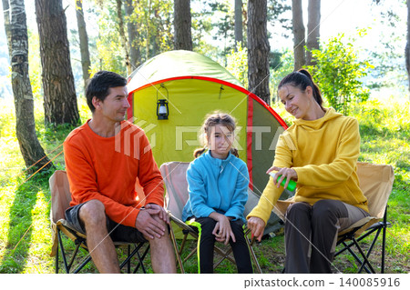 Mother and father apply bug spray to daughter, tent camping, outdoor picnic, protection from insect bites, safety priority. Mother and father apply bug spray to daughter, tent camping, outdoor picnic, protection from insect bites, safety priority. 140085916