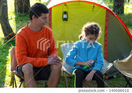 Daughter holding spray bottle, father watching, family outdoors at campsite, mosquito protection, bite allergy, summer health care, parent and child bonding. Daughter holding spray bottle, father watching, family outdoors at campsite, mosquito protection, bite allergy, summer health care, parent and child bonding. 140086018