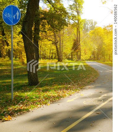 Blue round bicycle and pedestrian shared path sign on a metal pole next to an asphalt track winding through a beautiful public park filled with golden autumn trees. Blue round bicycle and pedestrian shared path sign on a metal pole next to an asphalt track winding through a beautiful public park filled with golden autumn trees. 140086562