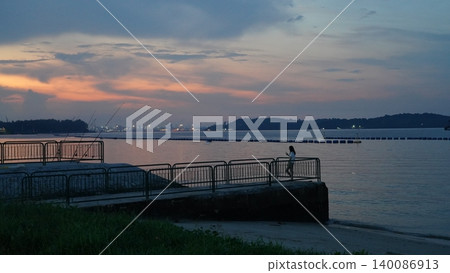 Woman on Pier at Dusk with Phone, Serene Coastal Sunset Woman on Pier at Dusk with Phone, Serene Coastal Sunset 140086913