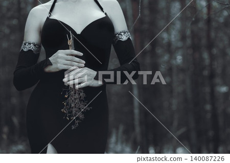 Faceless witch in gothic black dress and deathly pale skin holding dry plants in hands in dark spooky forest, copy space. Faceless witch in gothic black dress and deathly pale skin holding dry plants in hands in dark spooky forest, copy space. 140087226