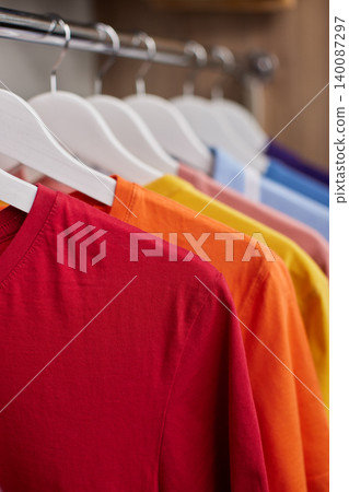 Colorful t-shirts and polo shirts hanging neatly on white hangers inside a wooden wardrobe, representing organization and selection Colorful t-shirts and polo shirts hanging neatly on white hangers inside a wooden wardrobe, representing organization and selection 140087297