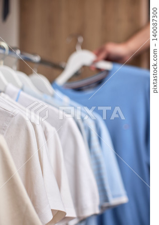 Hand selecting a blue t-shirt from a collection of clothes hanging on a rack in a closet or retail store Hand selecting a blue t-shirt from a collection of clothes hanging on a rack in a closet or retail store 140087300