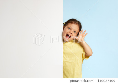 Happy little girl peeking from behind a white blank billboard, waving hand. Copyspace available for advertising message Happy little girl peeking from behind a white blank billboard, waving hand. Copyspace available for advertising message 140087309