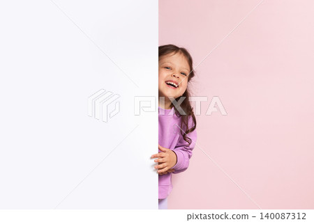 Smiling preschooler girl peeking from behind a large white empty banner, offering ample copy space for text or advertising Smiling preschooler girl peeking from behind a large white empty banner, offering ample copy space for text or advertising 140087312