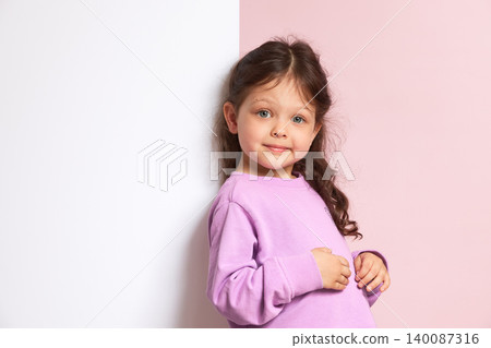 Young child with long brown hair wearing a purple sweater, standing against a pink and white background in a studio 140087316