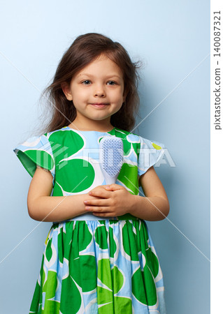 cute little child girl in a green and blue dress smiling, holding a light blue hairbrush on blue background 140087321