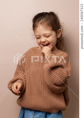 adorable little child girl smiling, biting finger, expressing joy, wearing brown knit sweater on beige background adorable little child girl smiling, biting finger, expressing joy, wearing brown knit sweater on beige background 140087323