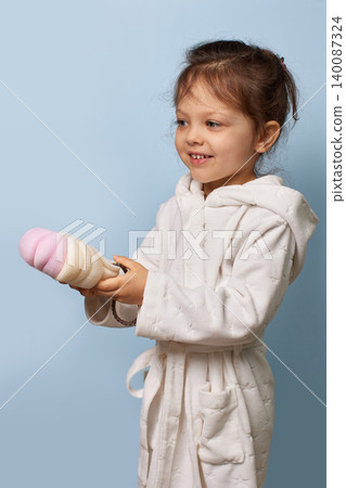 Little child girl smiling, wearing a white robe, holding a bath sponge for hygiene and play 140087324