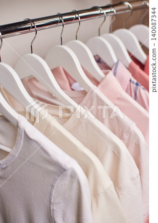 Row of various shirts hanging on white hangers on a metal rail inside a closet, showing organization and neatness 140087344