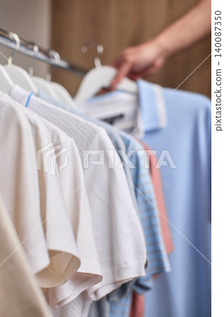 Hand selecting a blue t-shirt from a collection of clothes hanging on a rack in a closet or retail store Hand selecting a blue t-shirt from a collection of clothes hanging on a rack in a closet or retail store 140087350