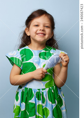adorable toddler girl smiling happily while holding a hairbrush on blue background, emphasizing childhood care 140087423