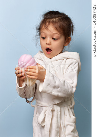 Little child girl in white bathrobe holding and looking at a pink bath sponge with wonder Little child girl in white bathrobe holding and looking at a pink bath sponge with wonder 140087428