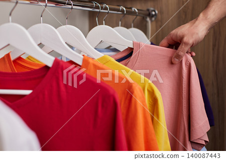 male hand selecting a pink short-sleeve t-shirt from a variety of colorful casual clothes organized on hangers in a wooden closet male hand selecting a pink short-sleeve t-shirt from a variety of colorful casual clothes organized on hangers in a wooden closet 140087443