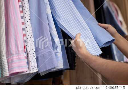 Hands browsing through a variety of men's shirts hanging on a rail inside a wooden closet, making a style choice Hands browsing through a variety of men's shirts hanging on a rail inside a wooden closet, making a style choice 140087445