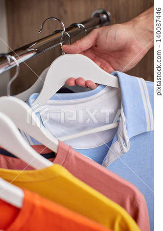male hand removing a light blue polo shirt from a clothes rail, surrounded by other hanging garments male hand removing a light blue polo shirt from a clothes rail, surrounded by other hanging garments 140087446