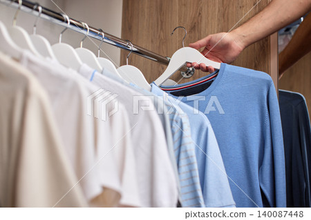 Hand selecting a light blue t-shirt with a white hanger from a selection of clothes on a rack in a wooden wardrobe 140087448