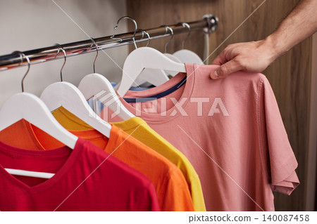 Man's hand selecting a pink short-sleeve t-shirt from a variety of colorful casual clothes organized on hangers in a wooden closet Man's hand selecting a pink short-sleeve t-shirt from a variety of colorful casual clothes organized on hangers in a wooden closet 140087458
